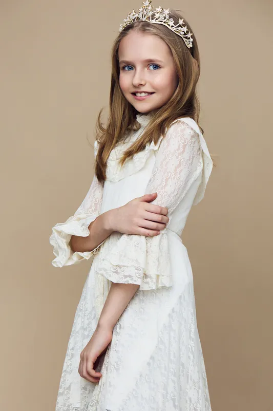 Young girl in elegant white dress with lace sleeves and a crown, smiling joyfully against a soft beige background, representing elegance and innocence