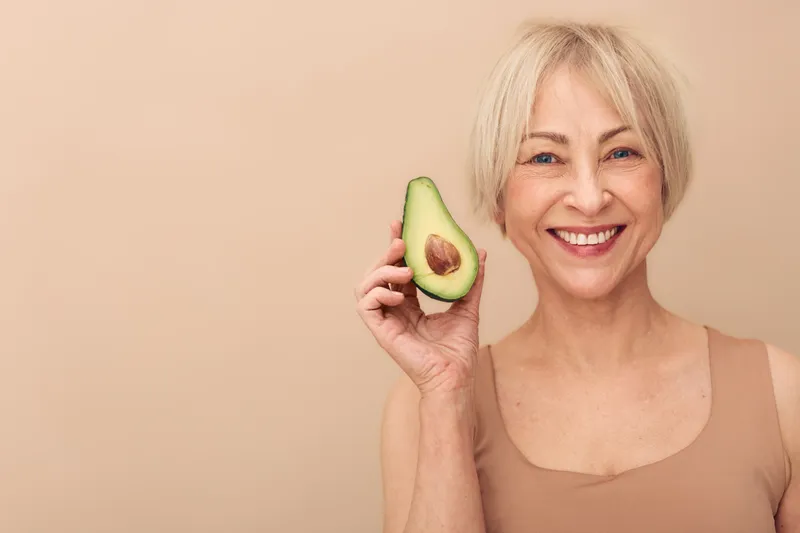 Smiling middle aged woman with short blonde hair holds half of ripe avocado near face. Nude background and casual tank top suggest natural beauty and healthy lifestyle concept.