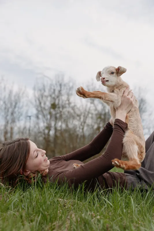 Woman lying on grass outdoors plays with a goat kid, capturing a cozy lifestyle moment of playful bonding and natural outdoors amid meadow and trees