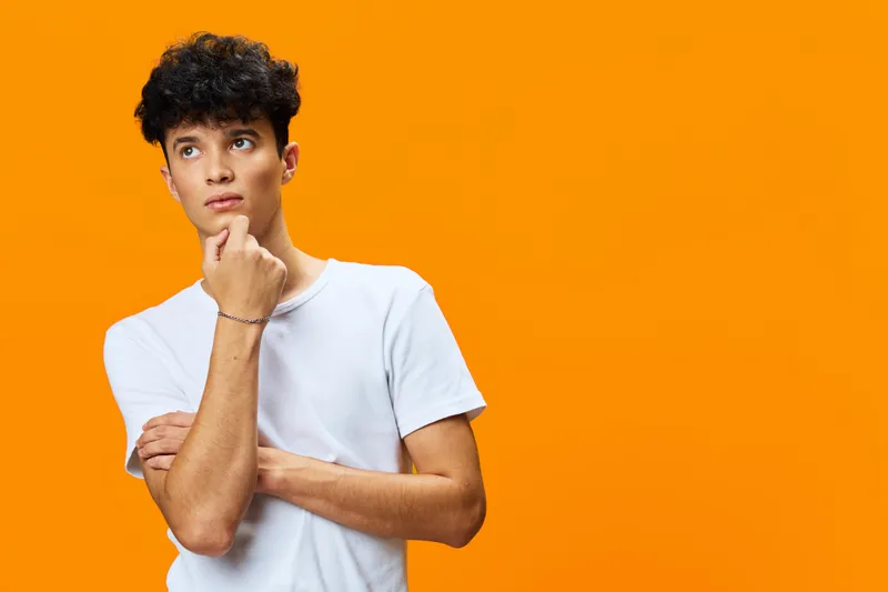 thoughtful young man in white t-shirt stands against bright orange background, looking pensive and curious with hand resting on chin in casual pose