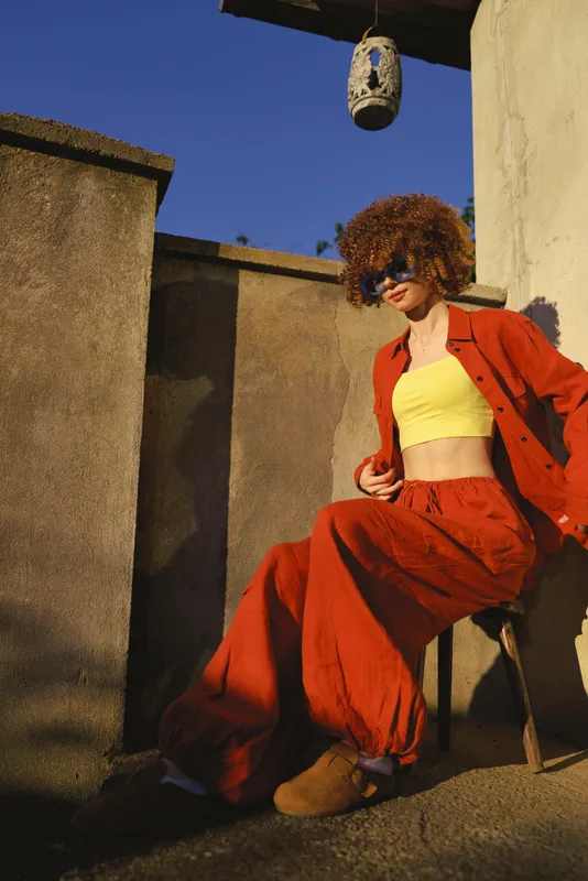 A woman in a bright red suit and yellow crop top sits on a balcony, stylish and relaxed, showcasing bold color styling for fashion lifestyle photography