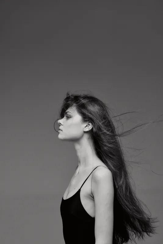 Black and white profile of a young woman with long flowing hair in a black dress against a plain gray background