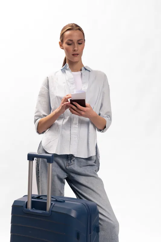 Young woman standing with a suitcase, holding a phone and looking at the screen. Travel concept with casual outfit and neutral background for vacation or business trip.