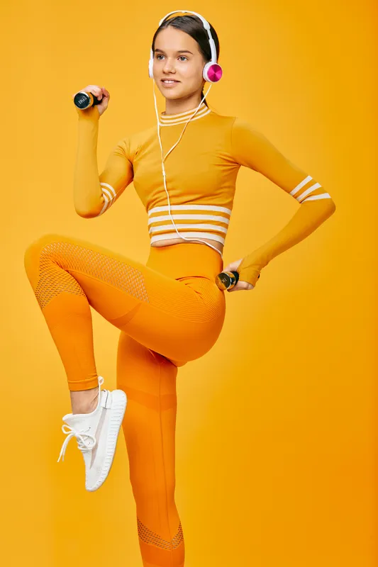 Woman fitness model performing a dynamic exercise with small dumbbells, wearing orange activewear, white sneakers and pink headphones against a vibrant orange studio background.