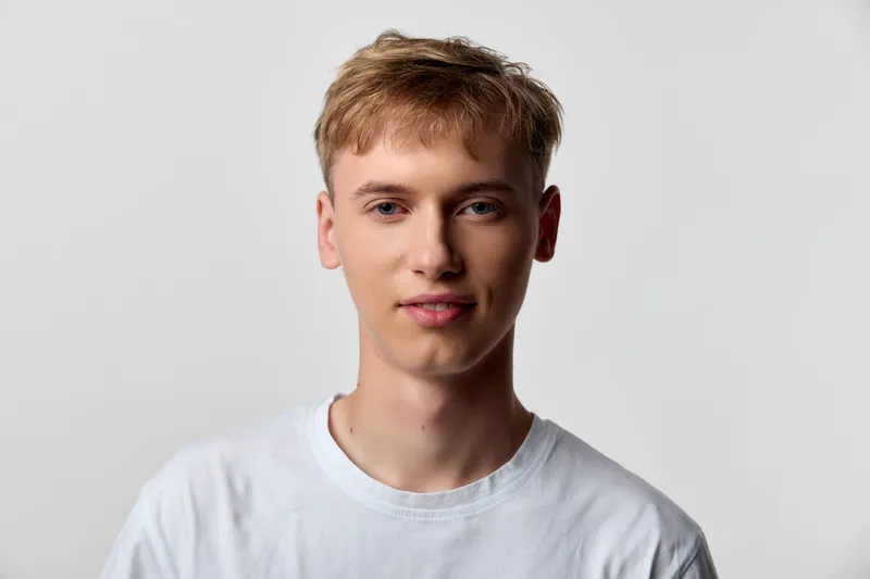 Young man in a white t shirt smiles softly against a plain background, ideal for lifestyle, fashion, or corporate projects, clean lighting and neutral tones for versatile advertising, editorial, or