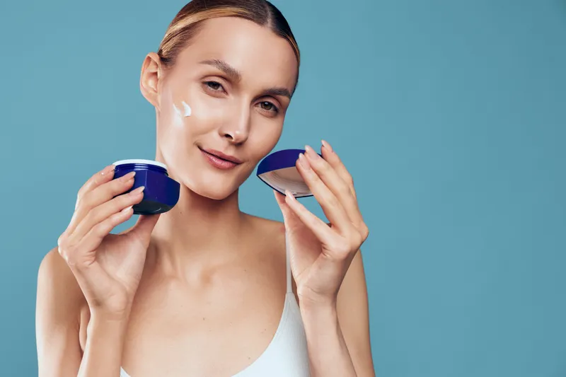 Young woman with clear skin applies moisturizing cream on her face while holding an open container against a plain blue background in a skincare routine.