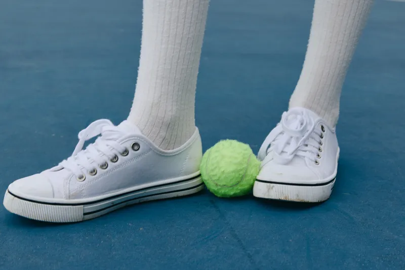 A young athlete in white sneakers stands on a blue court, with a bright green tennis ball nearby, capturing the spirit of youthful sports enthusiasm and competition