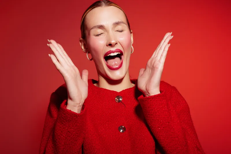 Excited woman in red sweater with closed eyes and open mouth expresses joy and surprise on red background. Concept of happiness, emotion, celebration, and positive energy.