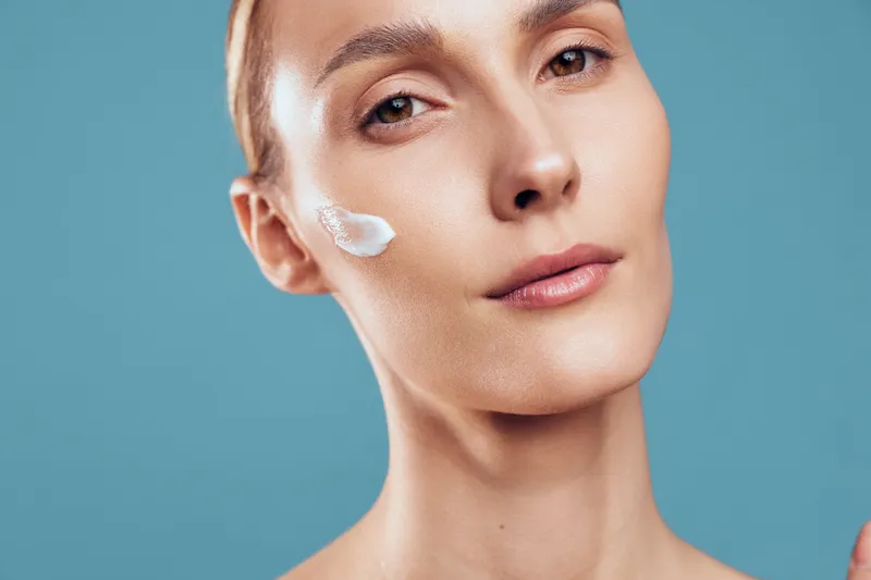 Closeup of woman with natural skin applying cream on cheek with calm expression isolated on blue background showing skincare routine and healthy face care concept.