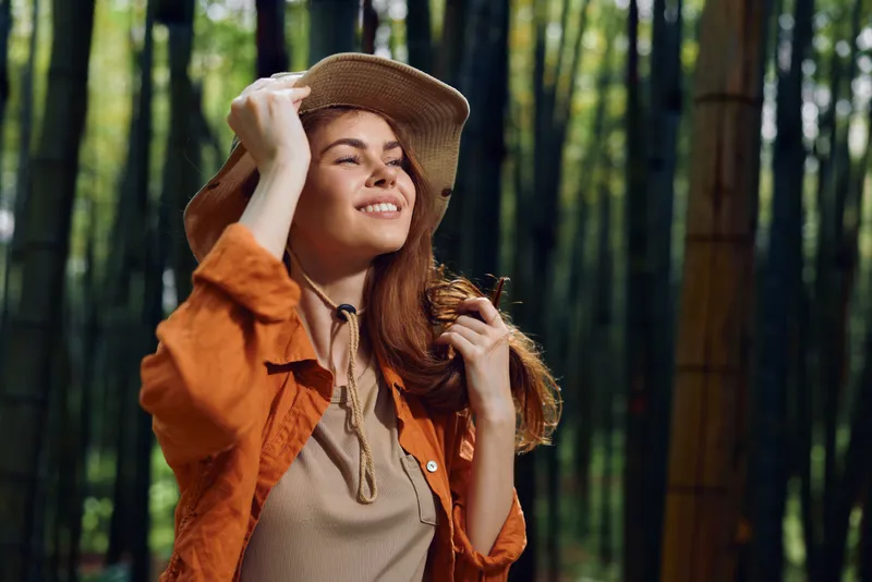 Woman with hat and smile stands in bamboo forest outdoors, portrait of happy traveler in orange jacket enjoying nature, sunlight and serene green foliage on a peaceful walk.