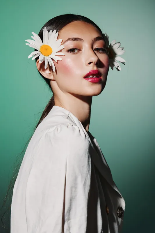 Portrait of a stylish woman with daisies in her hair wearing light clothes on green background. High fashion beauty concept with natural floral accessories and makeup.