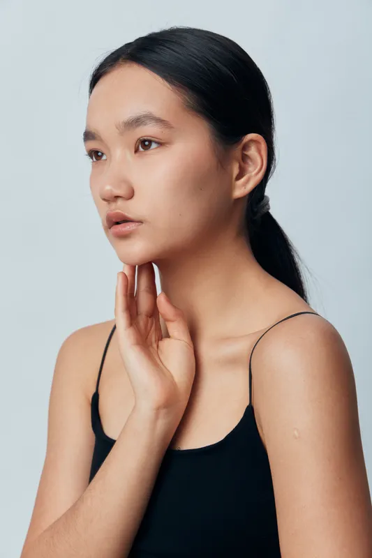 Young woman with smooth skin and black hair in ponytail wearing black tank top posing with thoughtful expression on light background. Beauty, fashion and natural portrait concept.