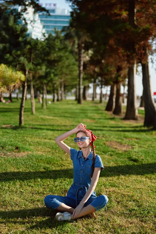 lifestyle woman in modern boho-western denim street style sitting cross-legged on grass with film color effect and sunglasses on a sunny day