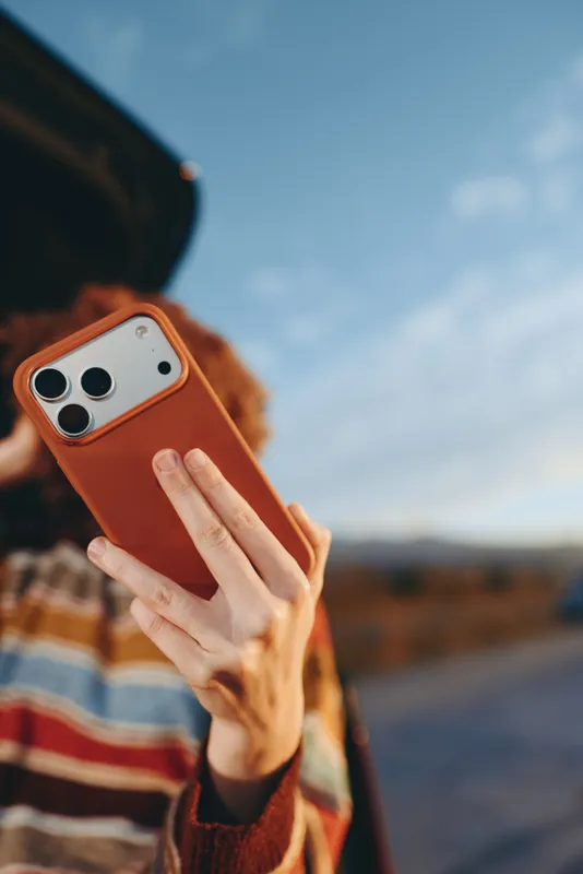 Woman lifestyle outdoors, closeup hands holding orange smartphone for selfie, striped sweater sleeves, blue sky and landscape background, daylight technology communication moment