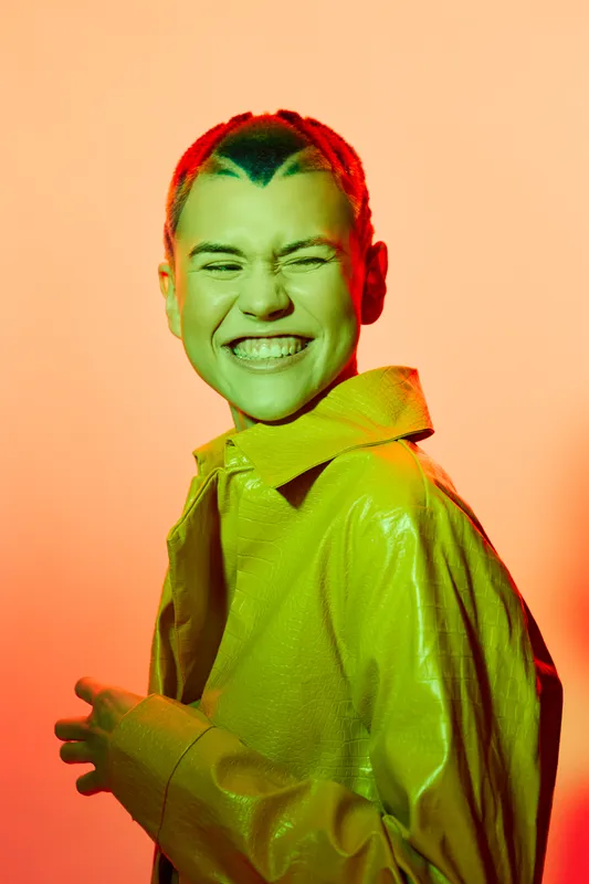 Smiling woman with short hair wearing a shiny green jacket, colorful vibrant lighting, joyful expression and warm orange background, modern fashion portrait.