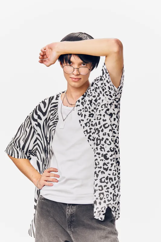 Young man wearing animal print shirt and glasses posing confidently with hand on forehead against white background in casual urban style.