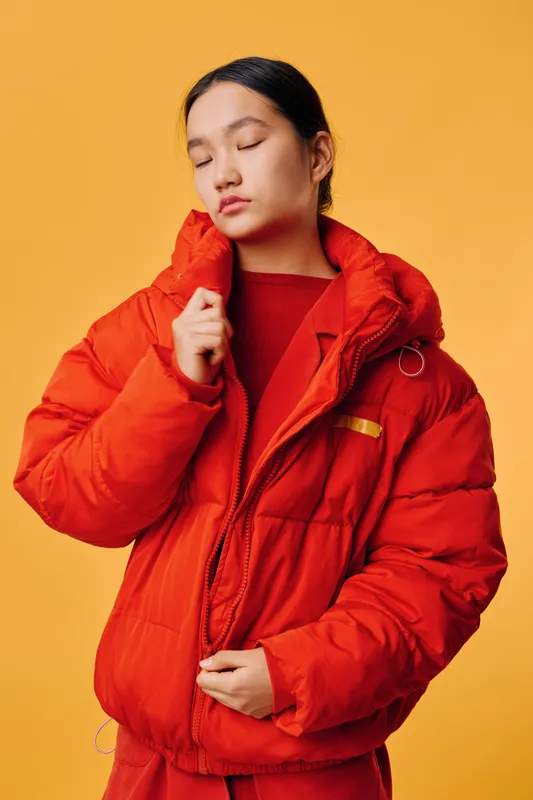 Woman wearing bright red puffer jacket and matching outfit posing with closed eyes on yellow background. Fashion, style, winter clothing and casual apparel with confident expression and studio
