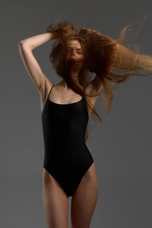 Woman with flowing hair wearing black bodysuit against gray background, dynamic motion, fashion style, modern elegance