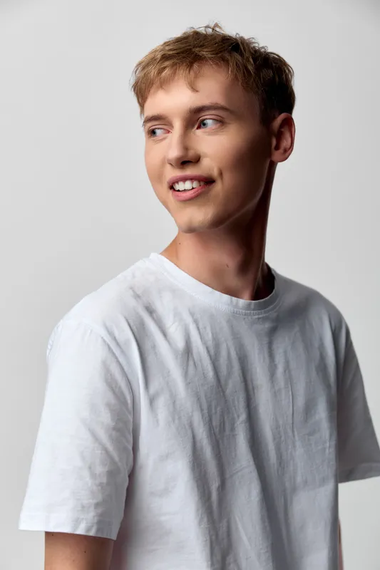 Young man in a white t shirt turned to the side, candid smile, studio lighting, casual fashion style, relaxed pose, clean background, friendly approachable vibe in studio shoot