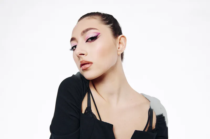Fashion model with creative bright purple eye makeup poses confidently wearing black off-shoulder top on white background. Editorial portrait highlighting beauty and style.