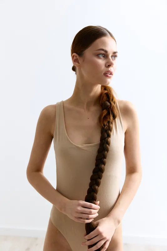 young woman with long braided hair wearing beige bodysuit standing against white background in natural light