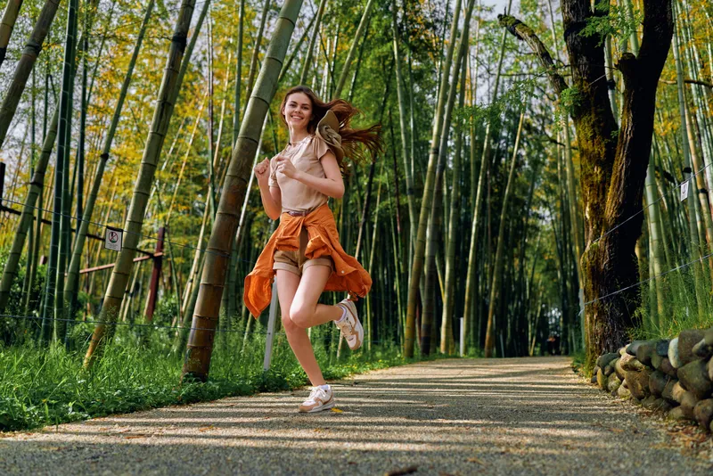 Woman running with a bright smile on a sunlit bamboo forest path, wearing summer skirt and sandals, joy and motion captured among tall green stalks and dappled light.