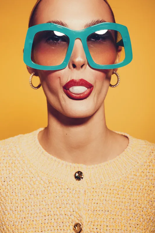 Close-up of a woman with oversized turquoise sunglasses and red lipstick making a playful face against a yellow background. Concept of fashion, fun, and bold style.