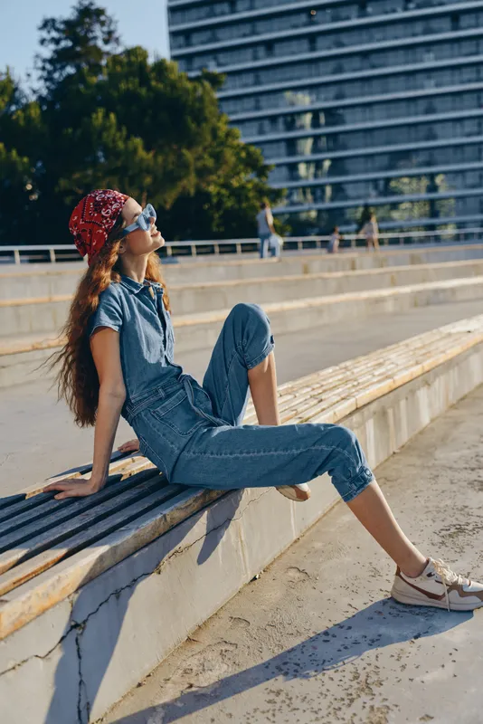 Lifestyle woman in modern boho-western denim street style relaxing outdoors with sunglasses and red bandana, enjoying sunny day in urban environment, film color tone.