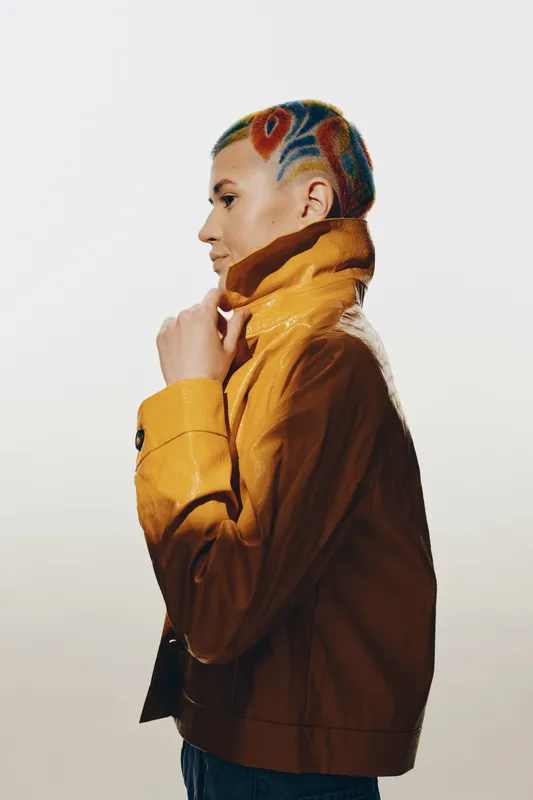 stylish woman with colorful patterned short hair wearing a mustard jacket standing sideways against white background in casual fashion pose