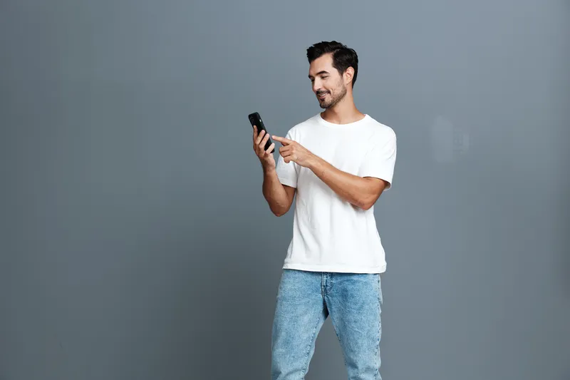 Smiling young man in casual white t-shirt stands against gray background, holding smartphone while pointing at screen, showcasing modern lifestyle and technology use.