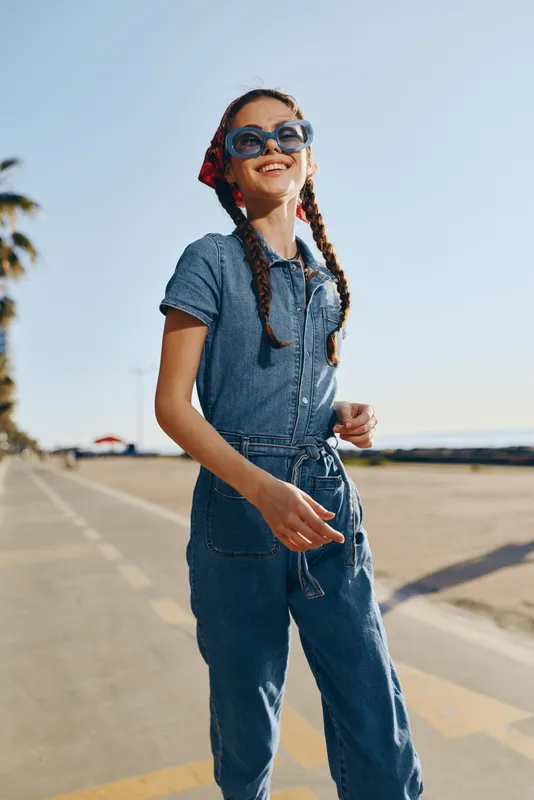 smiling girl in denim jumpsuit and sunglasses outdoors on a sunny day with blue sky, playful mood and carefree summer vibe