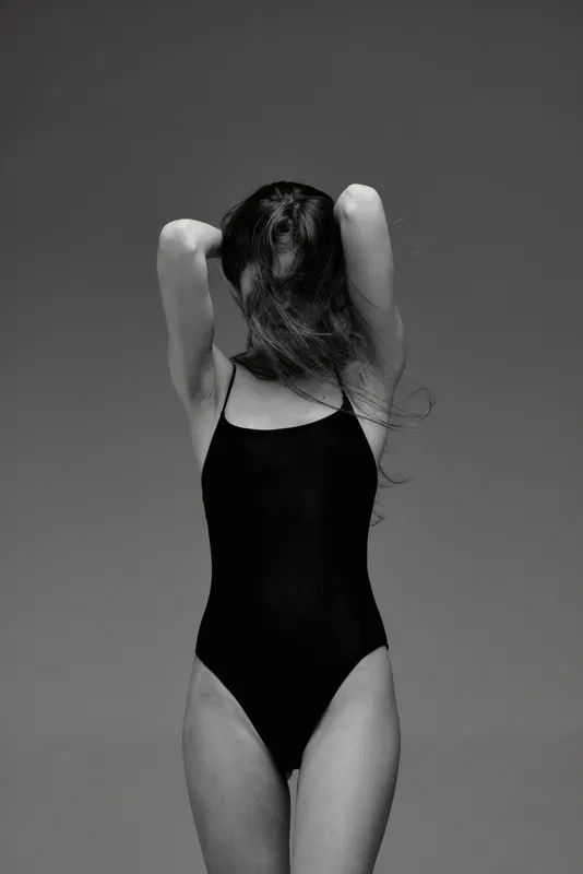 Black and white portrait of a woman in a black swimsuit with her hair covering her face, posing with arms raised.