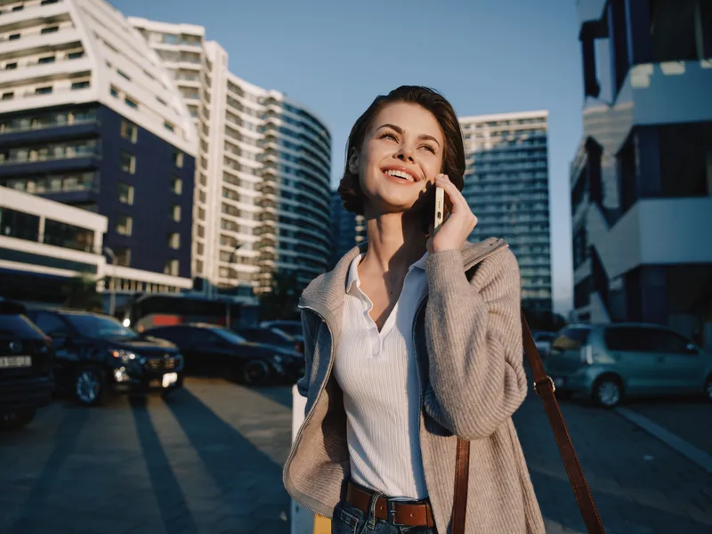 A cheerful businesswoman talks on her phone in a modern city street, wearing a blazer and white shirt, under bright sun, exuding confidence and a friendly, productive mood.