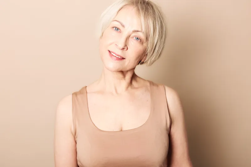 Smiling mature woman with short blonde hair wears beige tank top, posing in warm studio light on neutral background. Concept of natural beauty, aging gracefully, and confidence.
