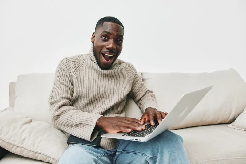 Young African man with a joyful expression using a laptop on a comfortable couch, wearing a cozy sweater, perfect for conveying remote work and digital communication themes