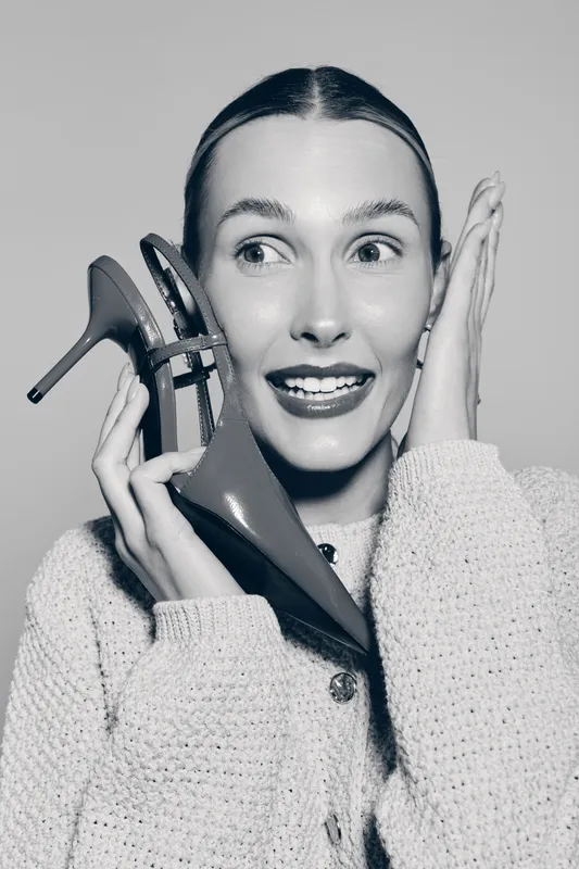 Young Caucasian woman with fair skin smiling and holding high heel shoes near face on grey background. Concept of fashion and beauty.
