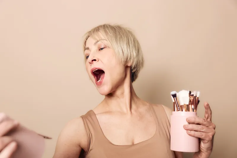 Middle aged woman with short blonde hair holding makeup brushes sings joyfully with open mouth on beige background. Concept for beauty routine, happiness, and self-expression.