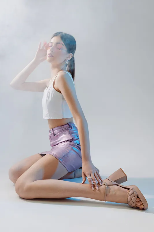 Young woman in a shiny skirt and white top poses kneeling with eyes closed and hand near face in soft light. Concept of beauty, fashion, and elegance in studio.