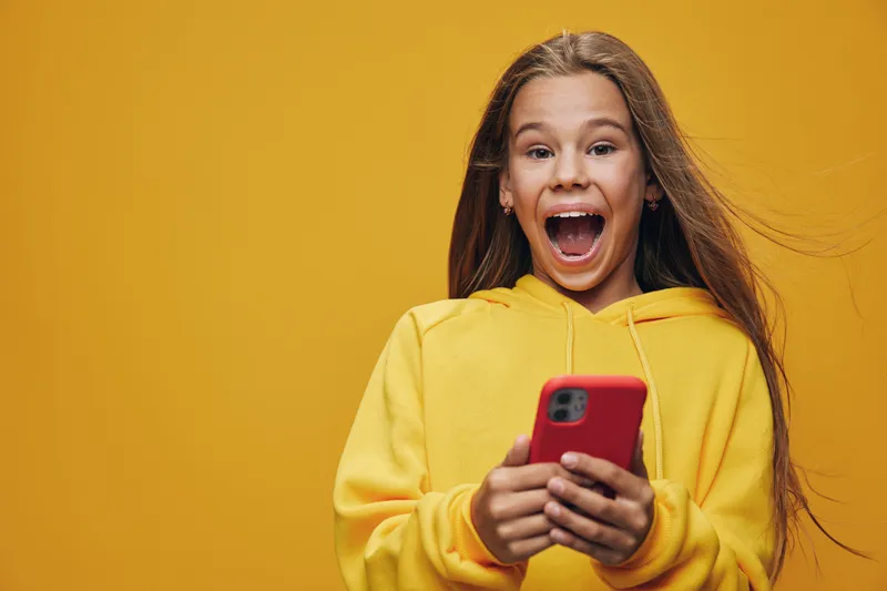 Girl with long hair in yellow hoodie looking excited and holding smartphone in hands on yellow background, happy child using mobile phone, communication and technology concept.