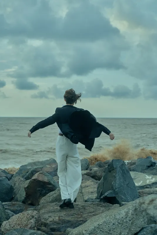 Man stands on a rocky shoreline facing the sea with wind lifting his jacket, calm yet determined mood, coastal landscape, horizon wide, solitary moment of resilience and contemplation amid crashing