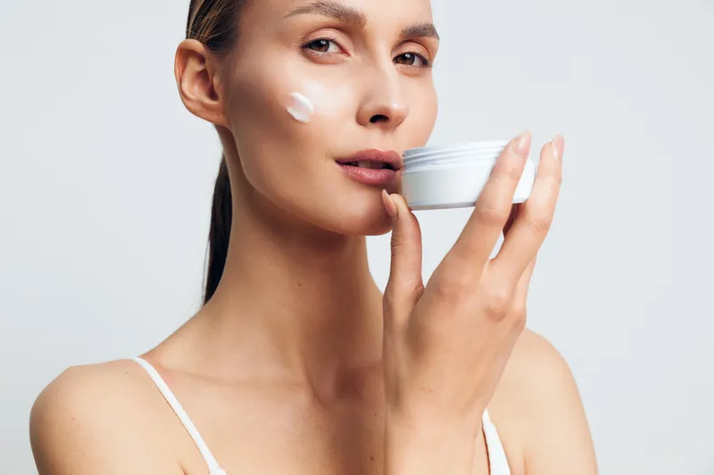 Young woman applies cream on face holding white jar close to skin with soft natural makeup isolated on light background for skincare beauty routine concept.