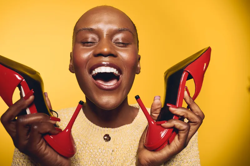 Joyful dark skinned African woman with bald head holds red high heels near face smiling with closed eyes on yellow background. Concept for fashion, happiness, and beauty expression.