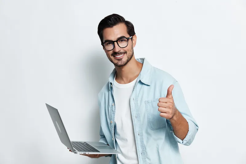 Smiling young man in glasses showing thumbs up while holding a laptop, isolated on light background. Positive emotions and modern lifestyle concept.