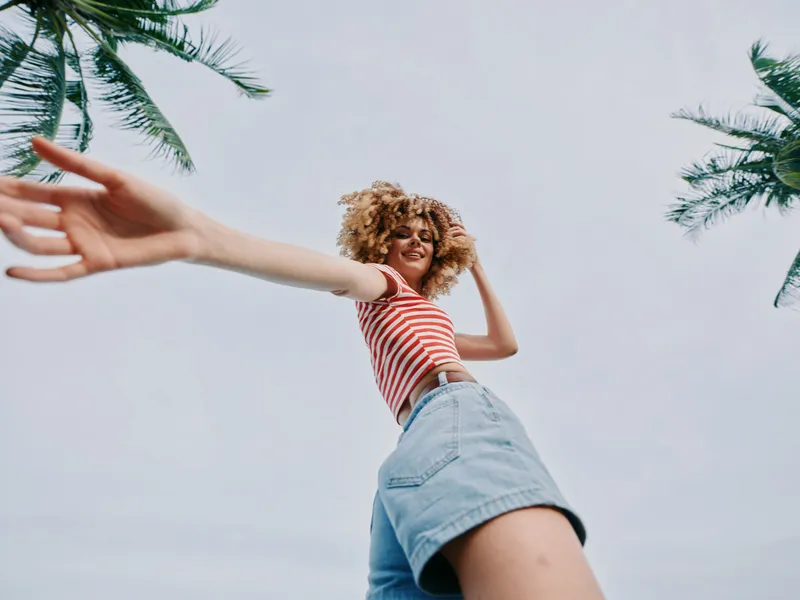 Low angle view of a joyful young woman in a red striped top and denim shorts, arms extended, sunny sky, palm trees, carefree summer mood, energetic outdoor pose.