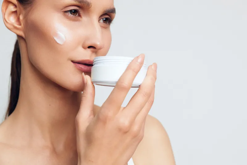 Closeup of a woman applying moisturizer on her cheek with a white cream jar in hand, smooth skin, natural beauty, soft lighting, minimalistic background, skincare routine concept