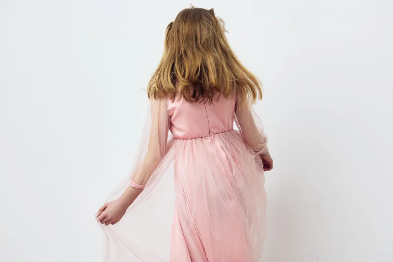 A young Caucasian girl in a pink tulle dress gracefully turns away, showcasing her long hair and delicate fabric against a minimalistic white background