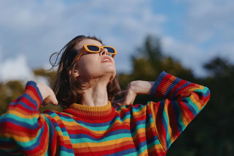 Young woman in her 20s, wearing a colorful striped sweater and yellow sunglasses, smiling and looking up while leaning back outdoors in a sunny park, casual lifestyle travel portrait.