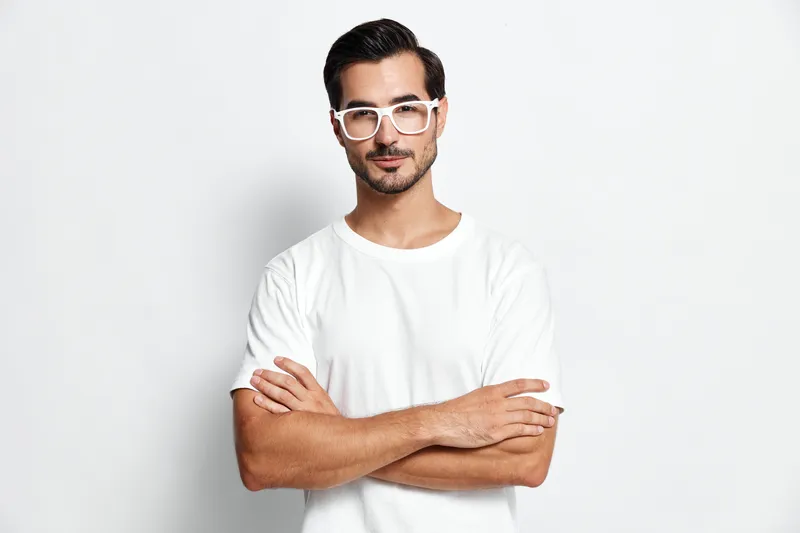 Confident young caucasian man in white t-shirt poses with arms crossed, stylish glasses and a thoughtful expression against a plain white background. Portrait concept.