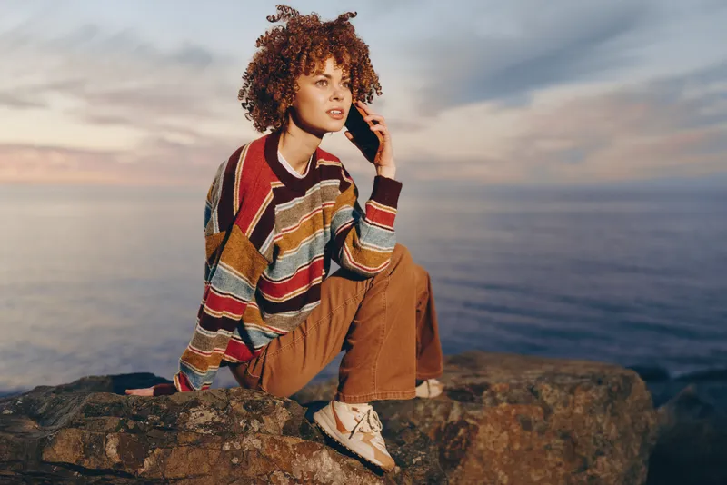 Woman lifestyle portrait outdoors, casual curly haired woman sits on cliff by ocean and sea rocks, wearing sweater poncho and jeans, sunset sky with clouds, relaxed travel vacation mood