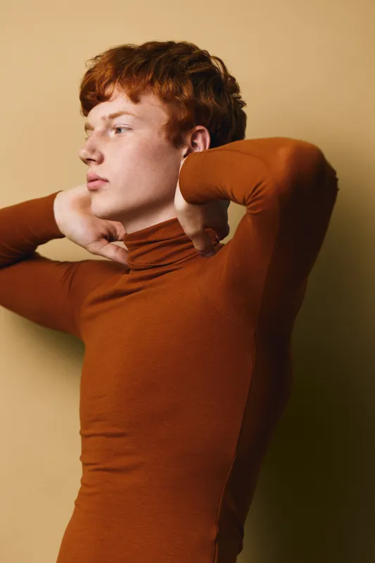Young man with red hair wearing brown turtleneck poses against beige background with hands behind neck and thoughtful expression in studio portrait.